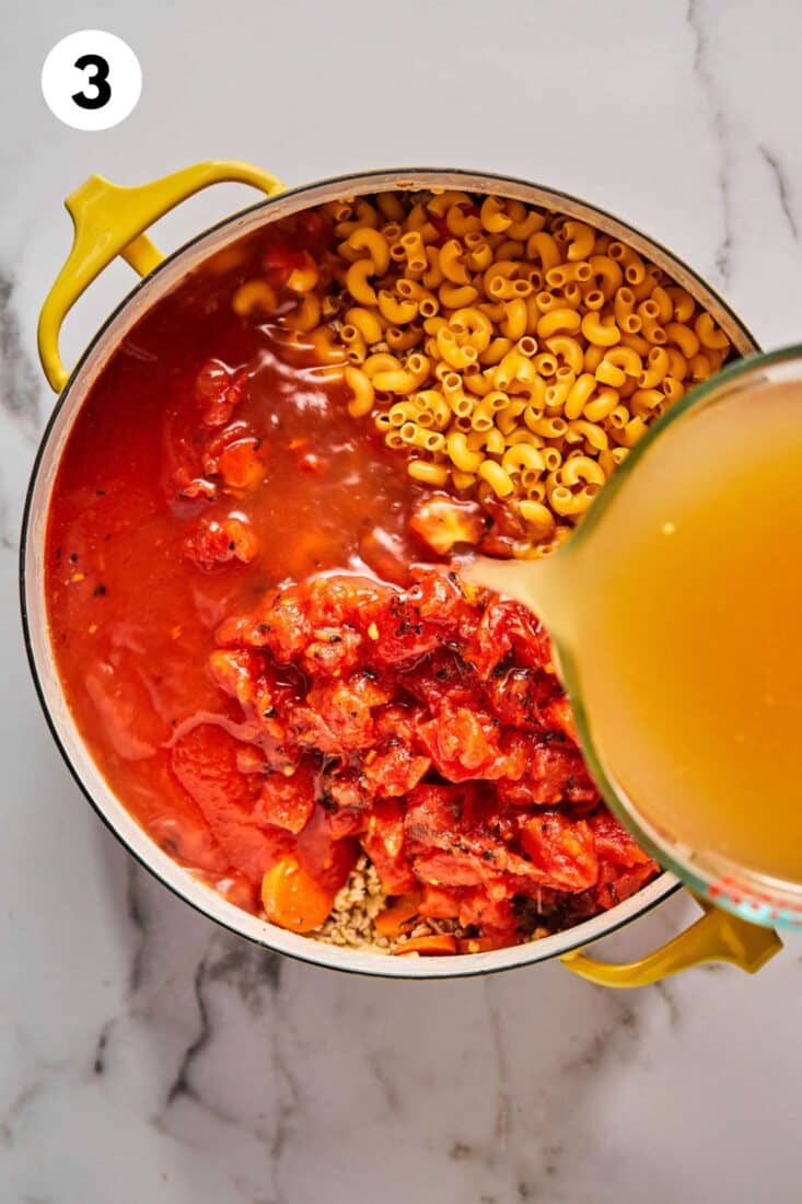 Pasta and tomatoes are added to the pot and broth is being added from a measuring cup.