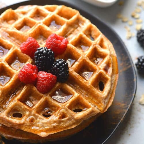 oatmeal waffle on a black plate with blackberries and raspberries on top and maple syrup.