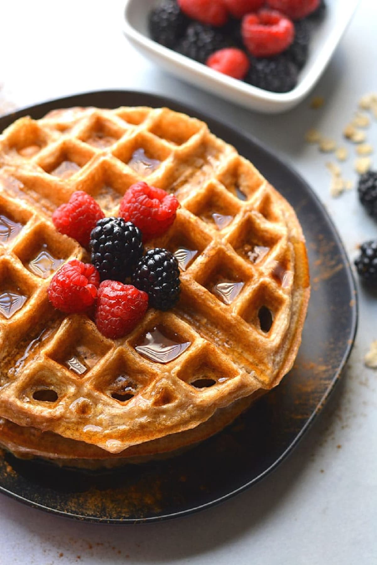 oatmeal waffle on a black plate with blackberries and raspberries on top and maple syrup.