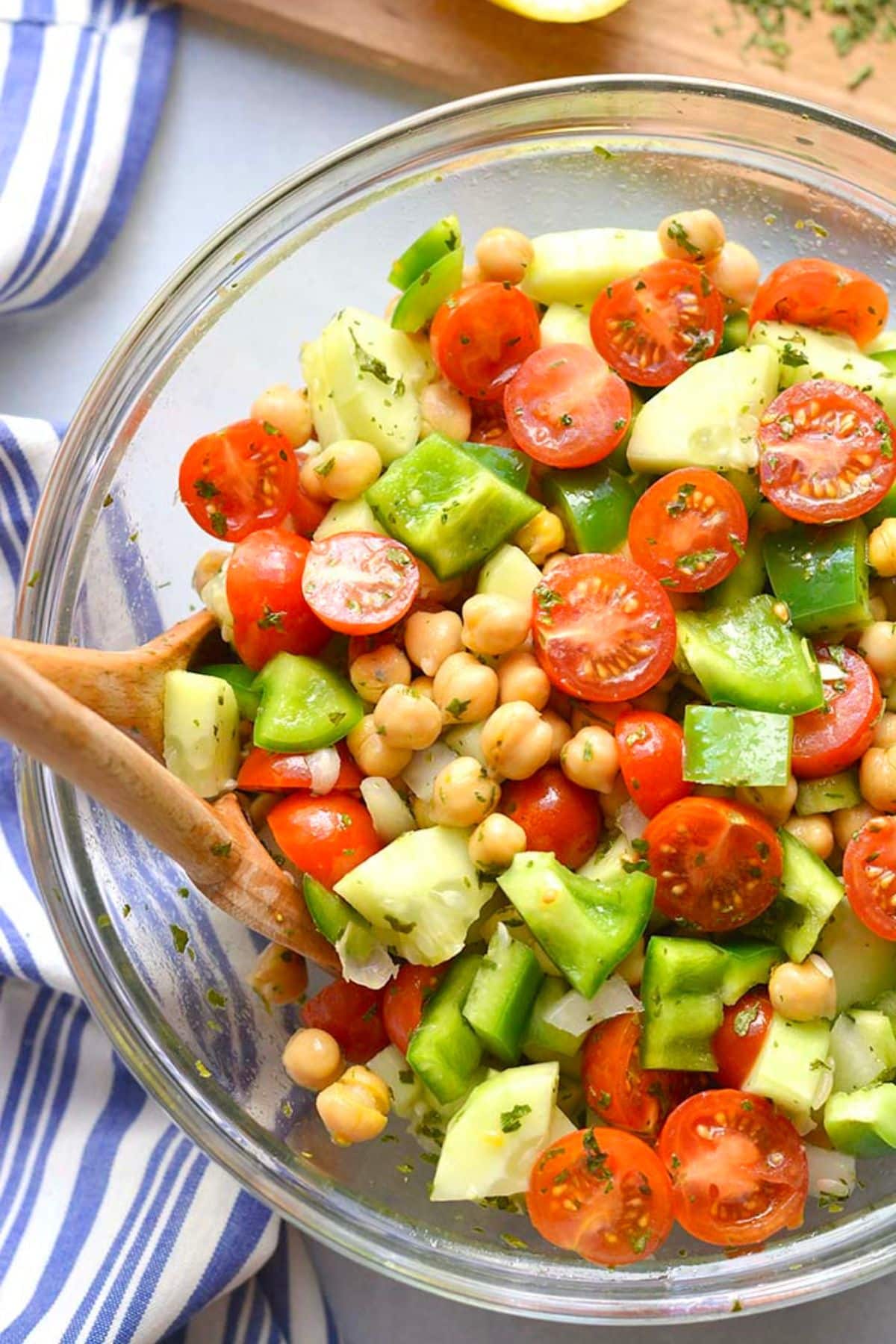A bowl of chickpea salad with tomato and cucumber and a wooden spoon resting in the bowl.