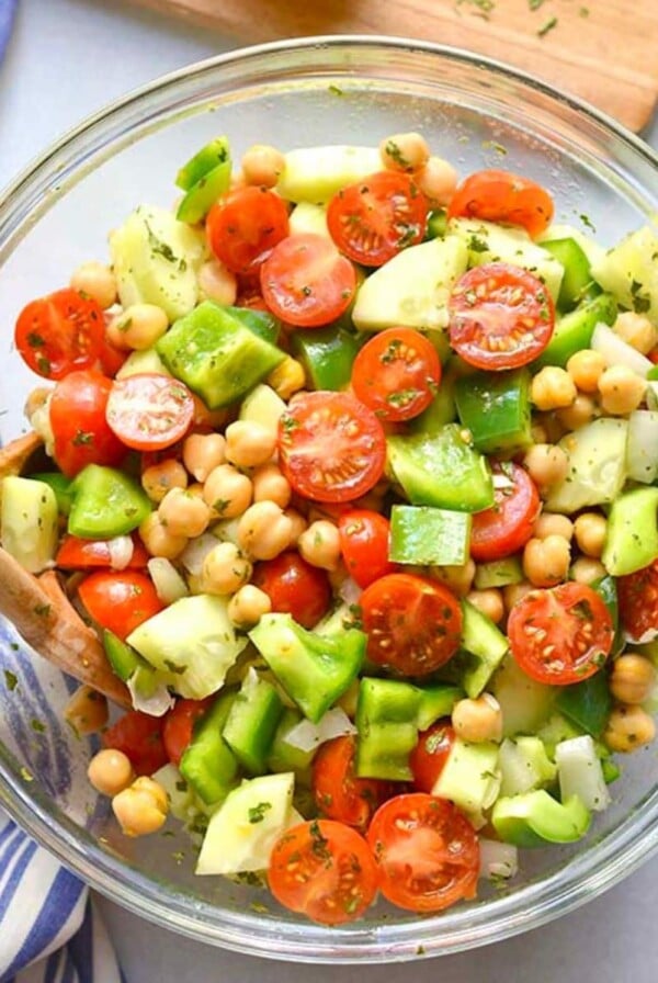 A bowl of chickpea tomato cucumber salad on a cutting board with a tea towel to the side.