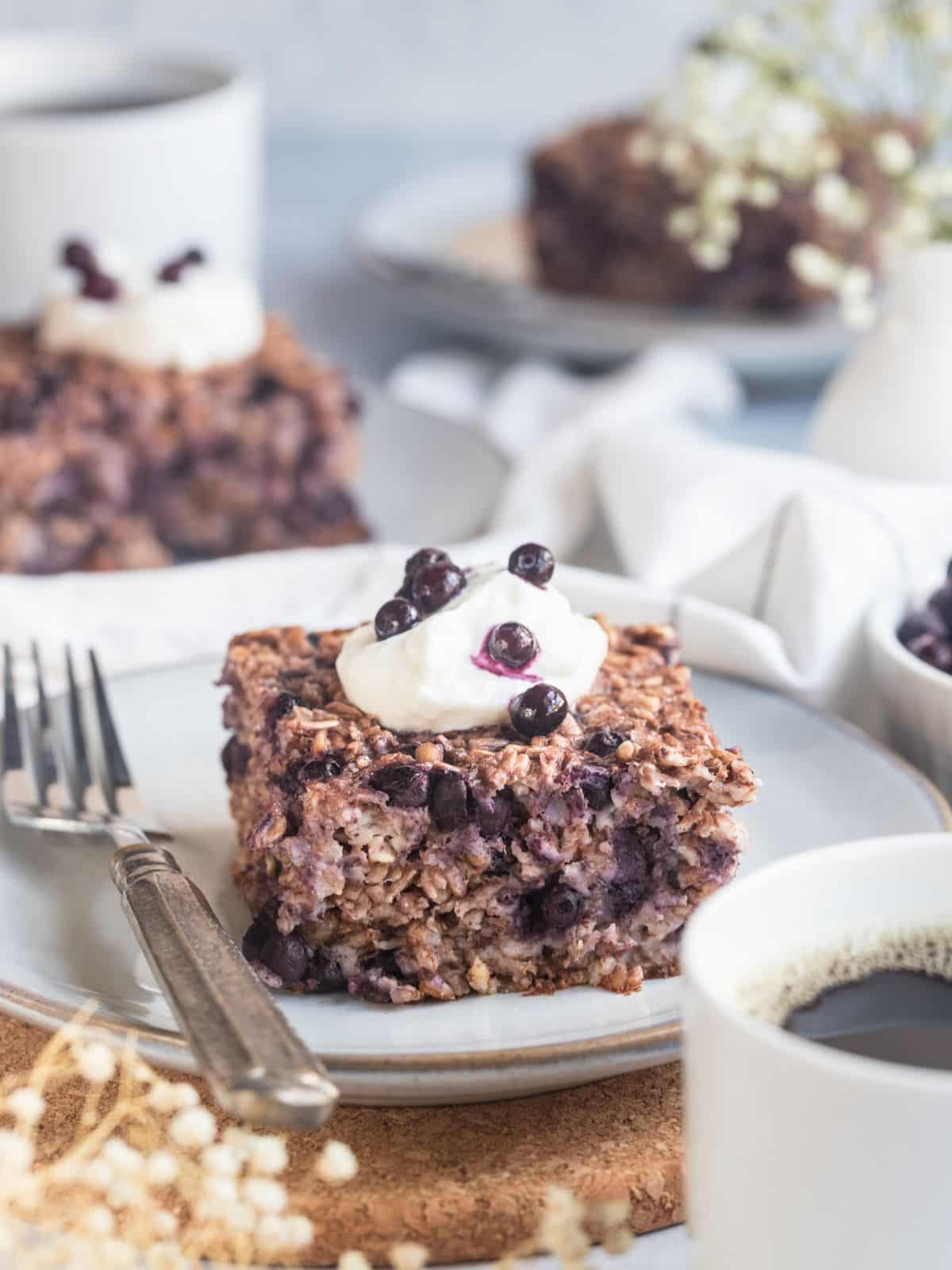 A square of healthy baked oatmeal on a plate topped with a dollop of yogurt and some blueberries. 