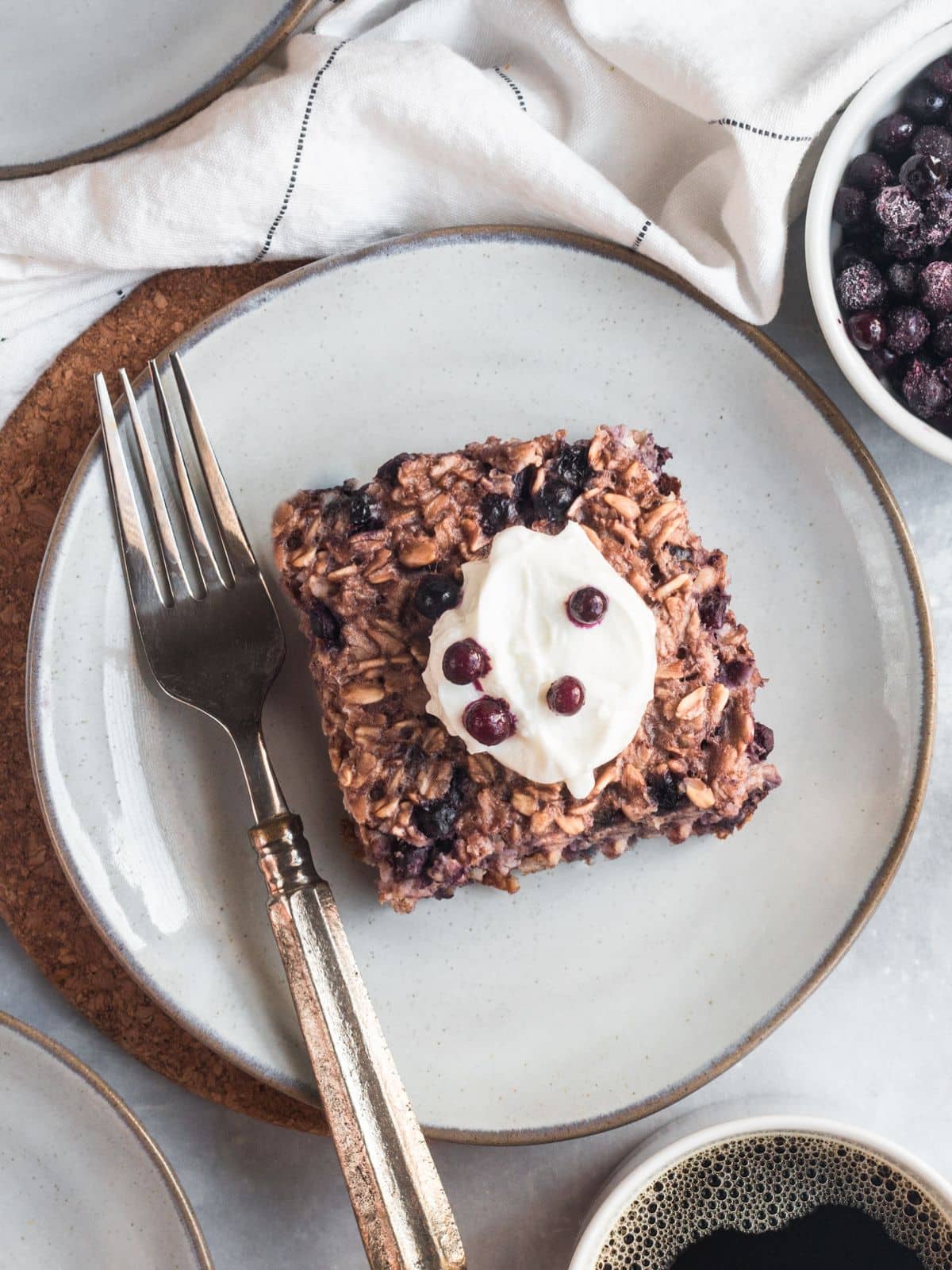 A square of healthy baked oatmeal on a plate topped with some greek yogurt and blueberries with a fork to the side. 