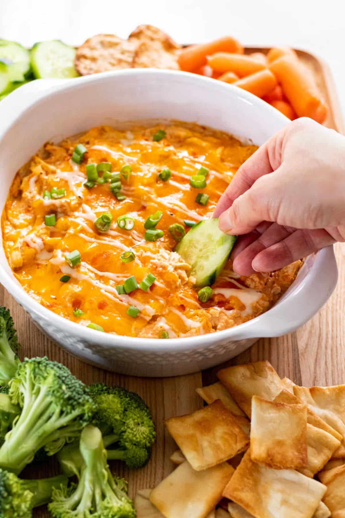 A round white casserole dish with healthy buffalo wing dip with a hand dipping into it with a cucumber.