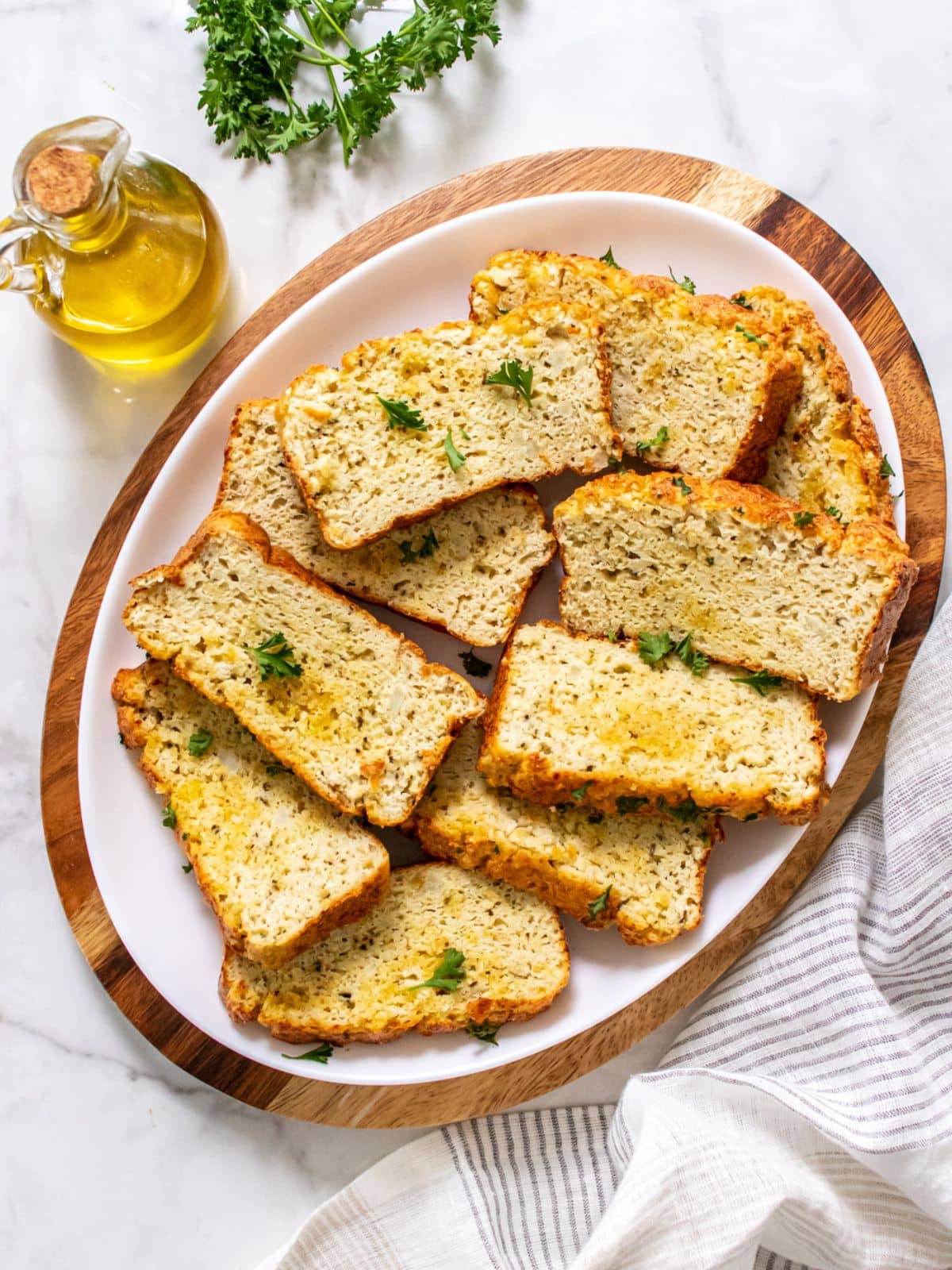 A platte of cauliflower bread on a wooden board next to a bottle of olive oil and parsley.