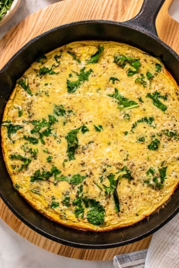 Kale frittata cooked in a cast iron skillet on a wooden cutting board.