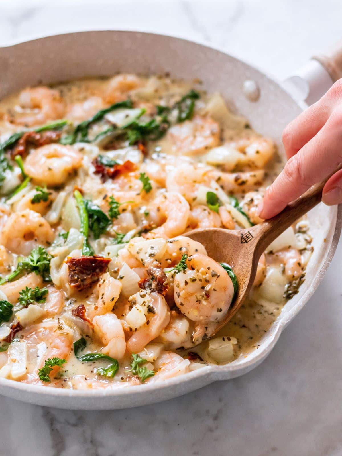 Tuscan shrimp recipe made in a skillet with a hand holding a wooden spoon in it.