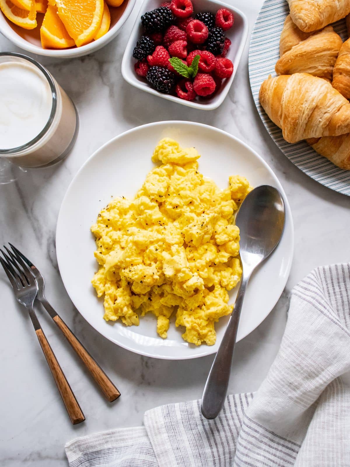 A plate of Greek yogurt scrambled eggs on the table with croisssants, a bowl of berries, and orange slices.