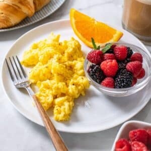 Greek yogurt scrambled eggs served up on a plate wiht a bowl of berries and a slice of orange.