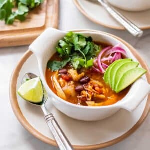 A bowl of healthy chicken tortilla soup on the table with limes, onion, cilantro, and avocado.