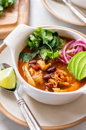 A bowl of healthy chicken tortilla soup on the table with limes, onion, cilantro, and avocado.