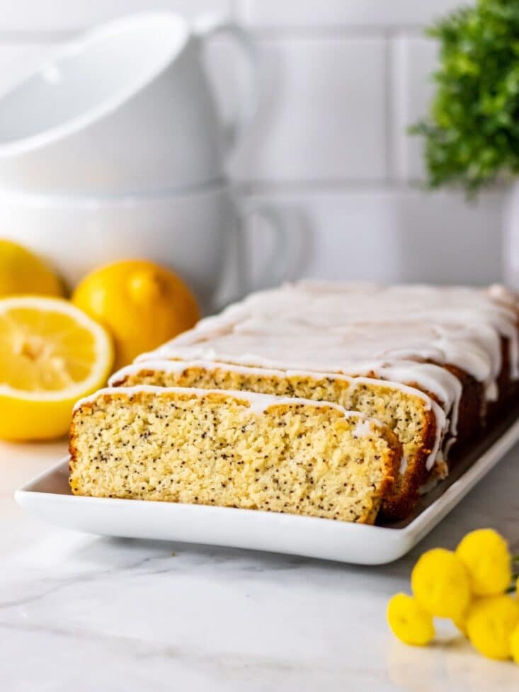 Sliced, frosted lemon and poppyseed loaf on a white plate with lemons and lemon peel artfully scattered around.