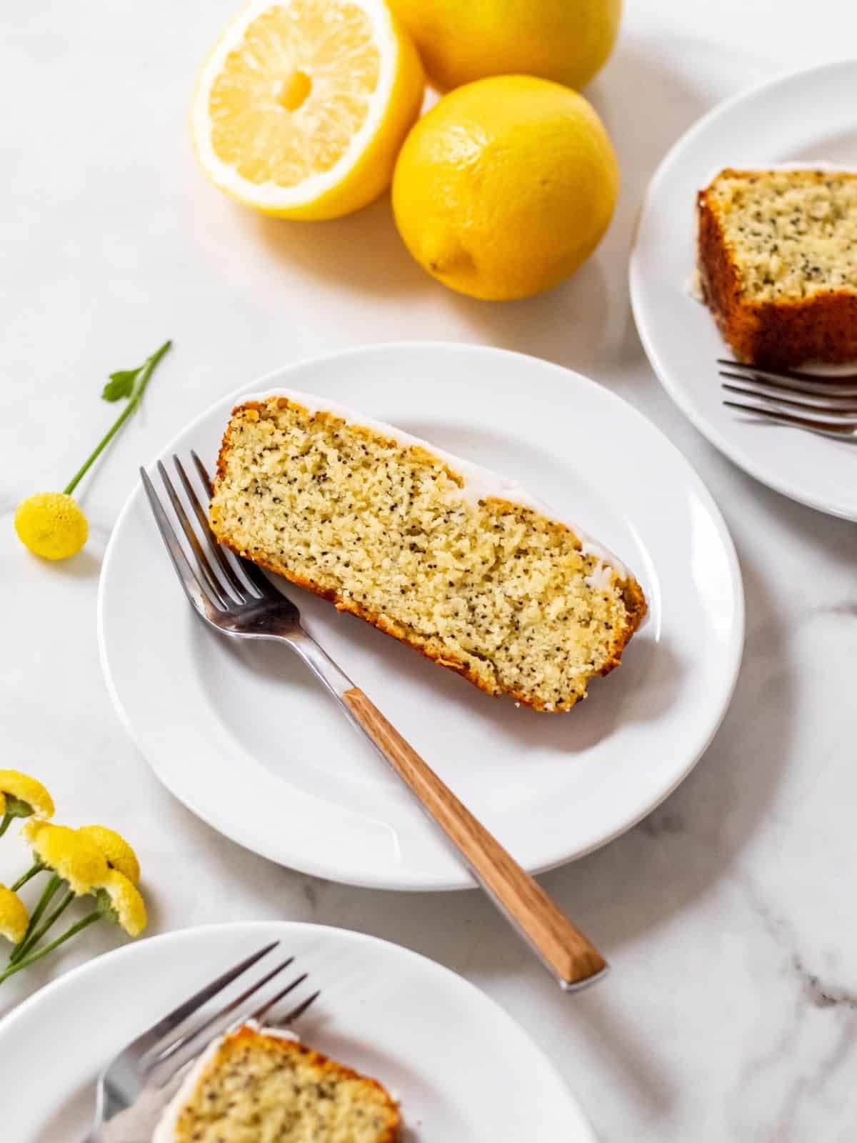 A slice of frosted lemon and poppyseed on a white plate, with a fork resting nearby. 