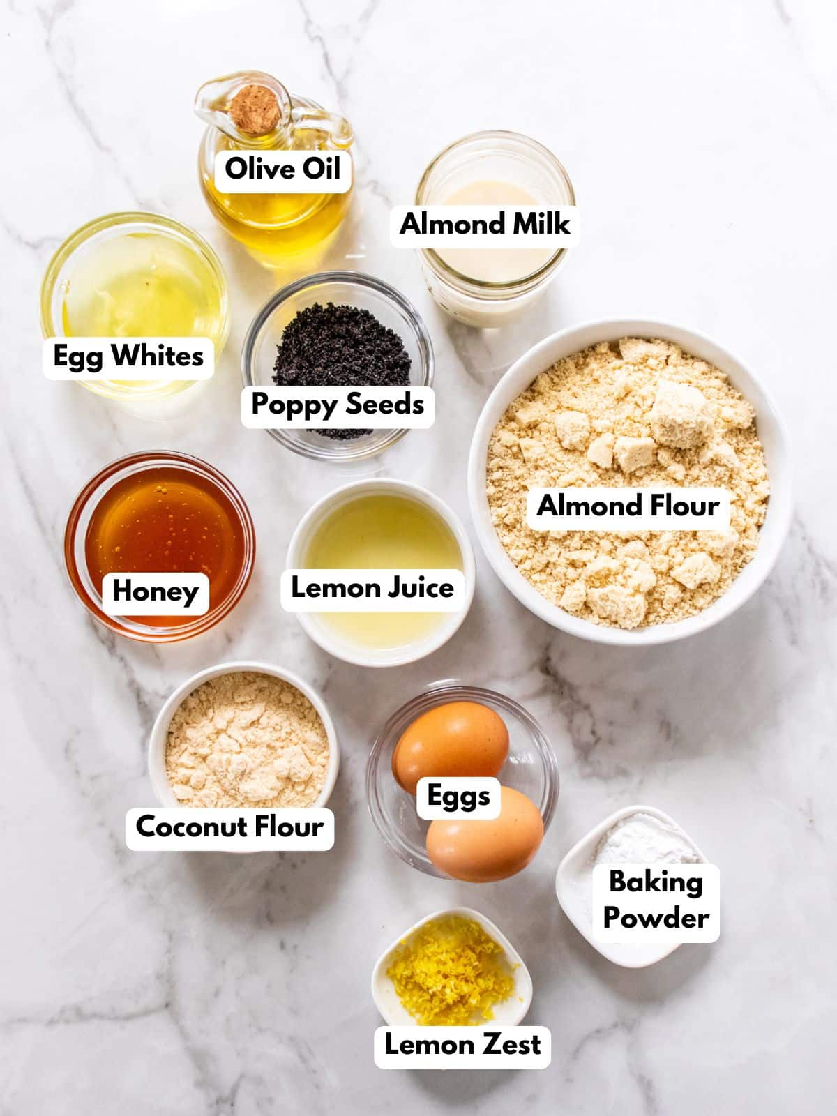Ingredients for healthy lemon cake labeled, in bowls, on a white marbled countertop.