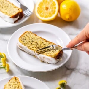 A hand with a fork, cutting into a slice of healthy lemon loaf cake sitting on a white plate.