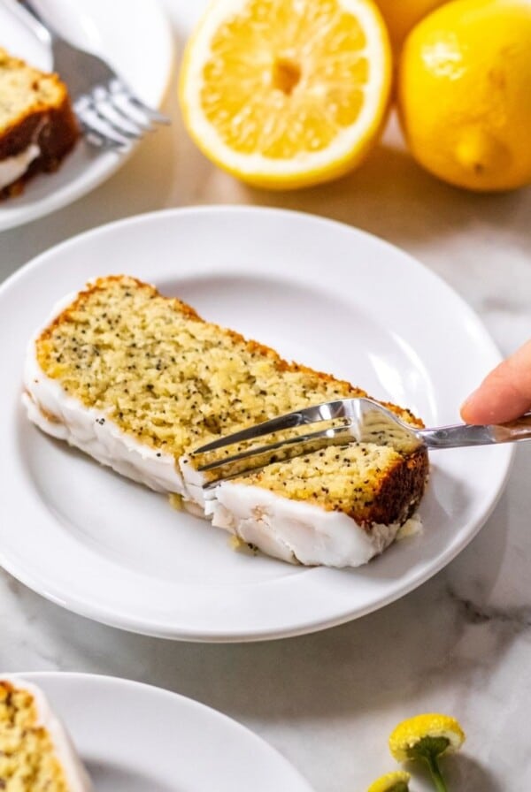 A hand with a fork, cutting into a slice of healthy lemon loaf cake sitting on a white plate.