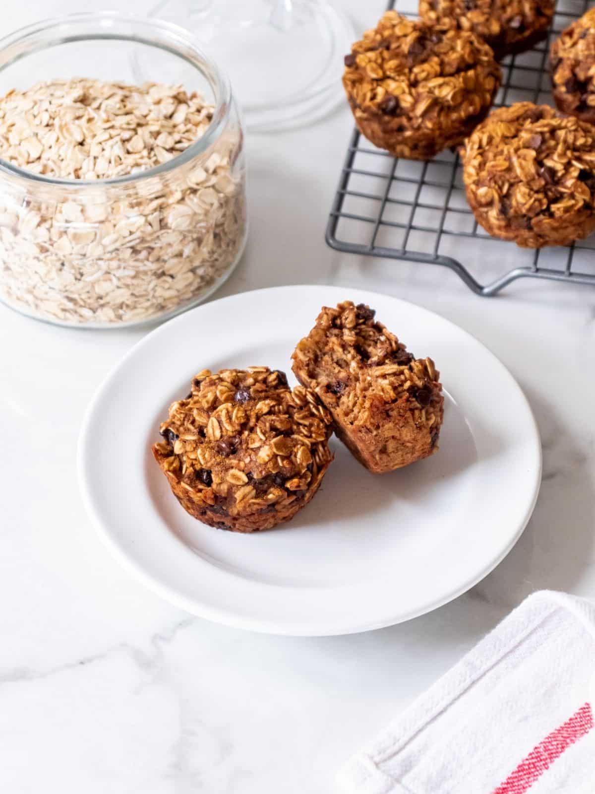 Two banana and peanut butter muffins on a plate with a jar of oats and more muffins on a cooling rack in the background.