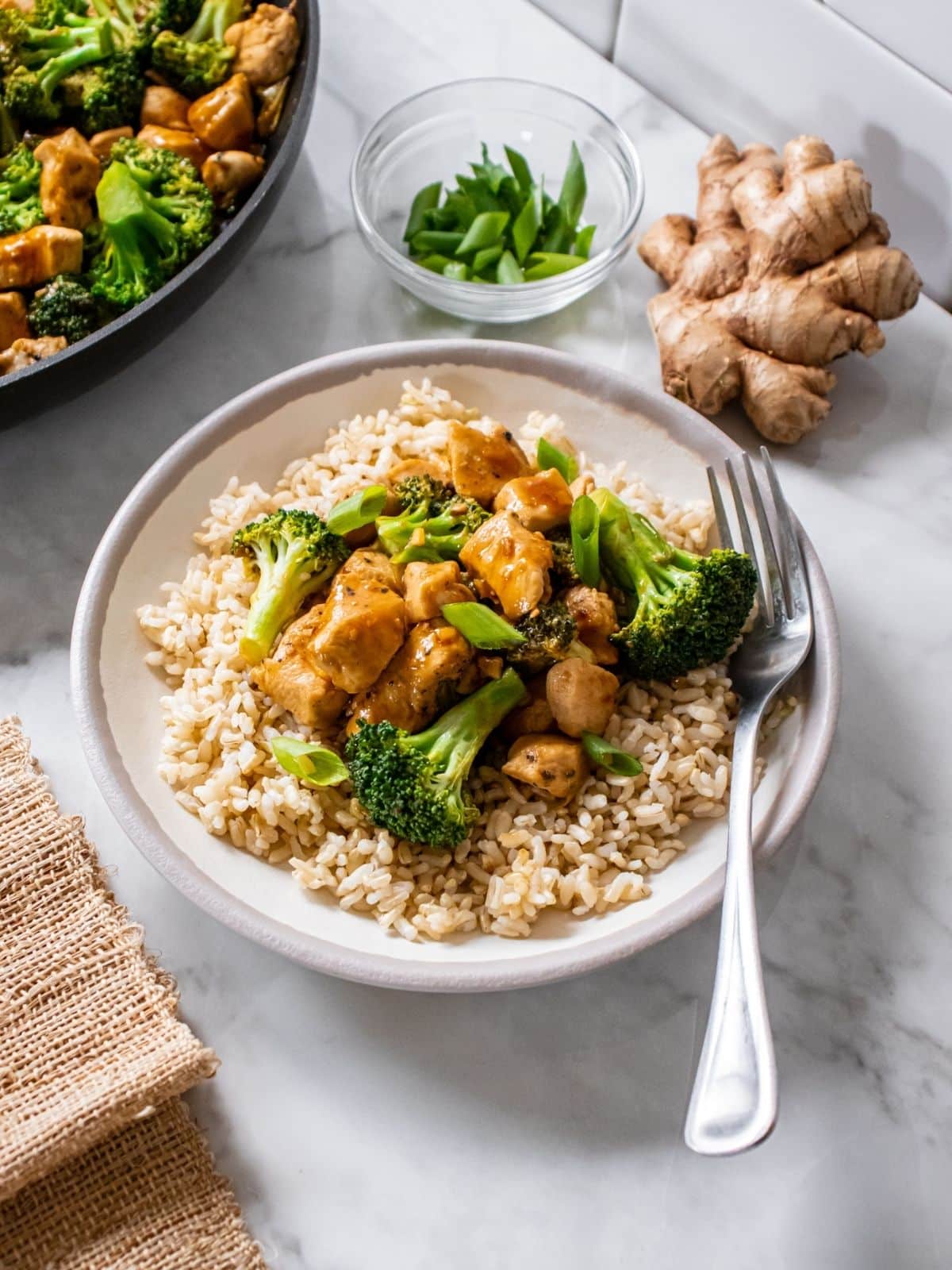A white bowl with rice, chicken, and broccoli with a fork resting on the side sits on a marbled countertop, with a chunk of fresh ginger and a small bowl of scallions to behind.