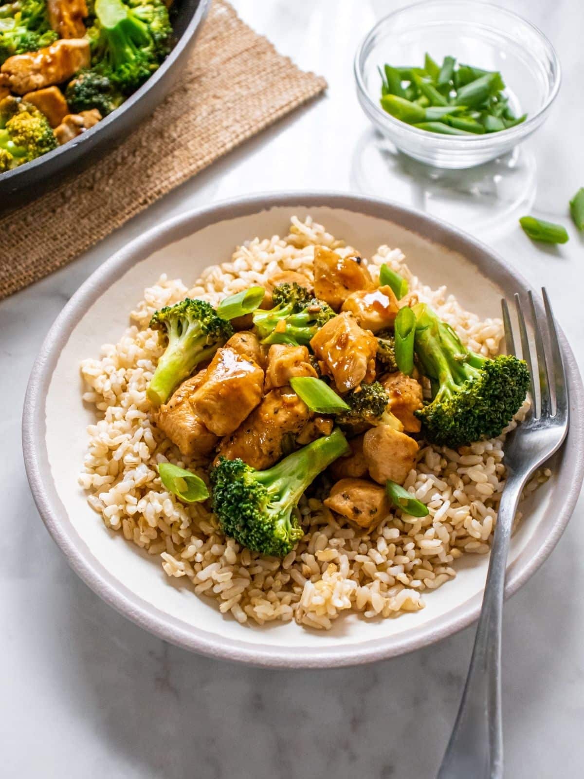 Chicken and broccoli on a bed of rice served in a white bowl with a fork resting on the side, and a small glass dish of scallions at the back.