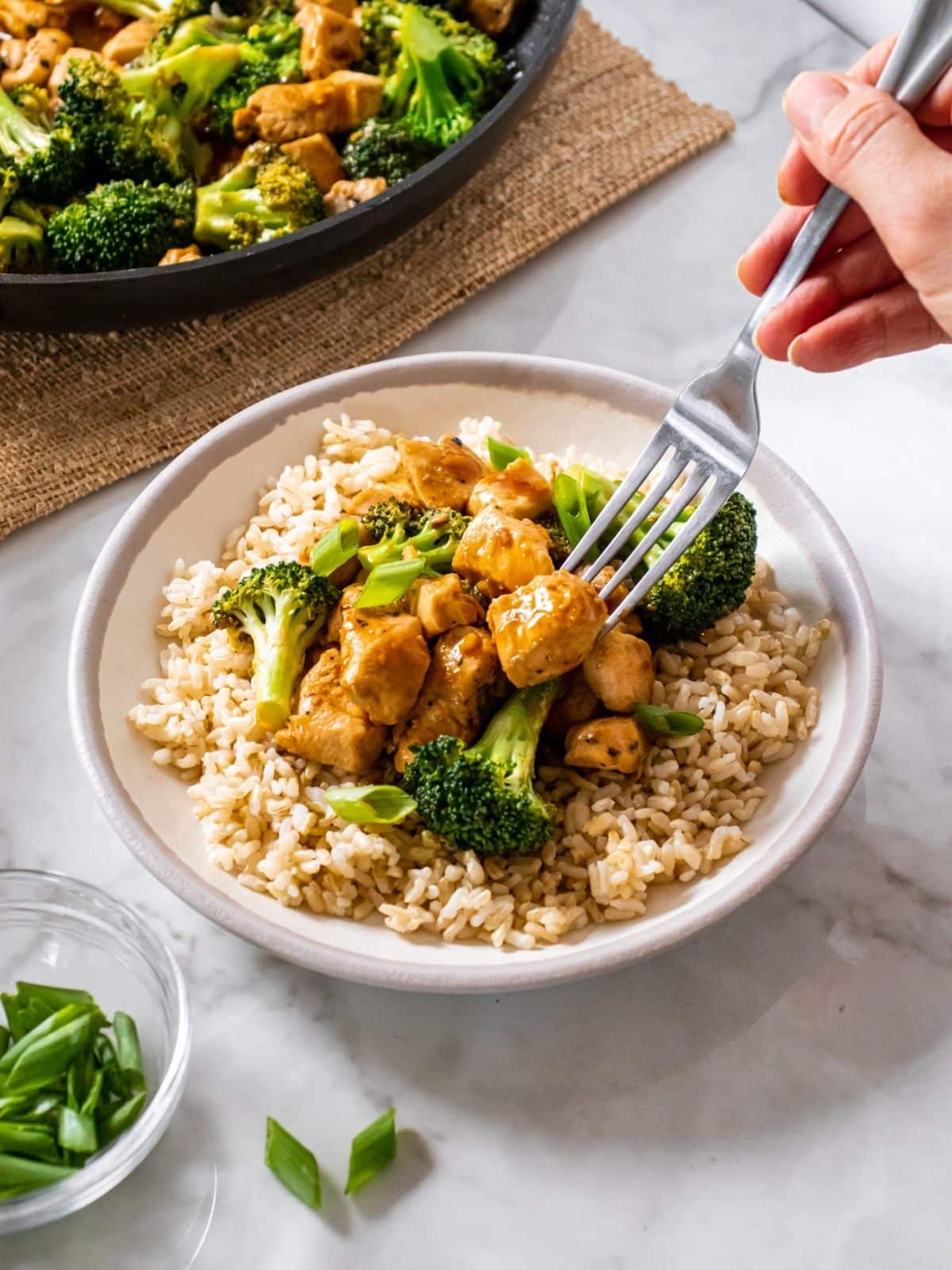 Chicken and broccoli stir fry over rice in a bowl, with a hand holding a fork, about to dig in. 
