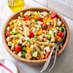 A bowl of healthy Greek pasta salad on a counter with a jar of olive oil behind it and spoons in the bowl.