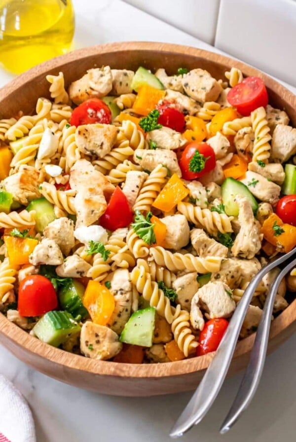 A bowl of healthy Greek pasta salad on a counter with a jar of olive oil behind it and spoons in the bowl.