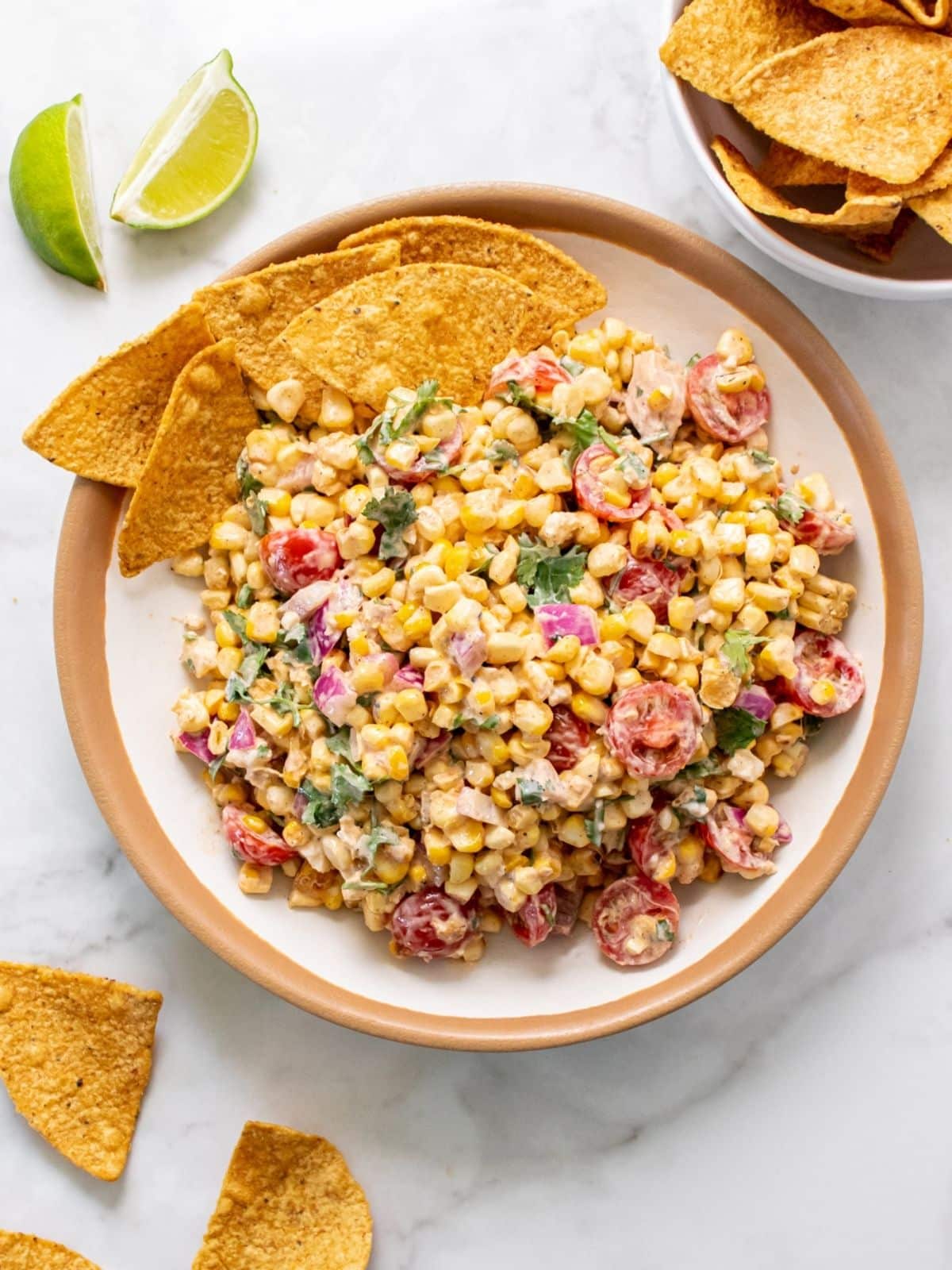 Healthy Mexican street corn served in a brown and cream bowl with tortilla chips on the side, with lime wedges and more chips scattered around.
