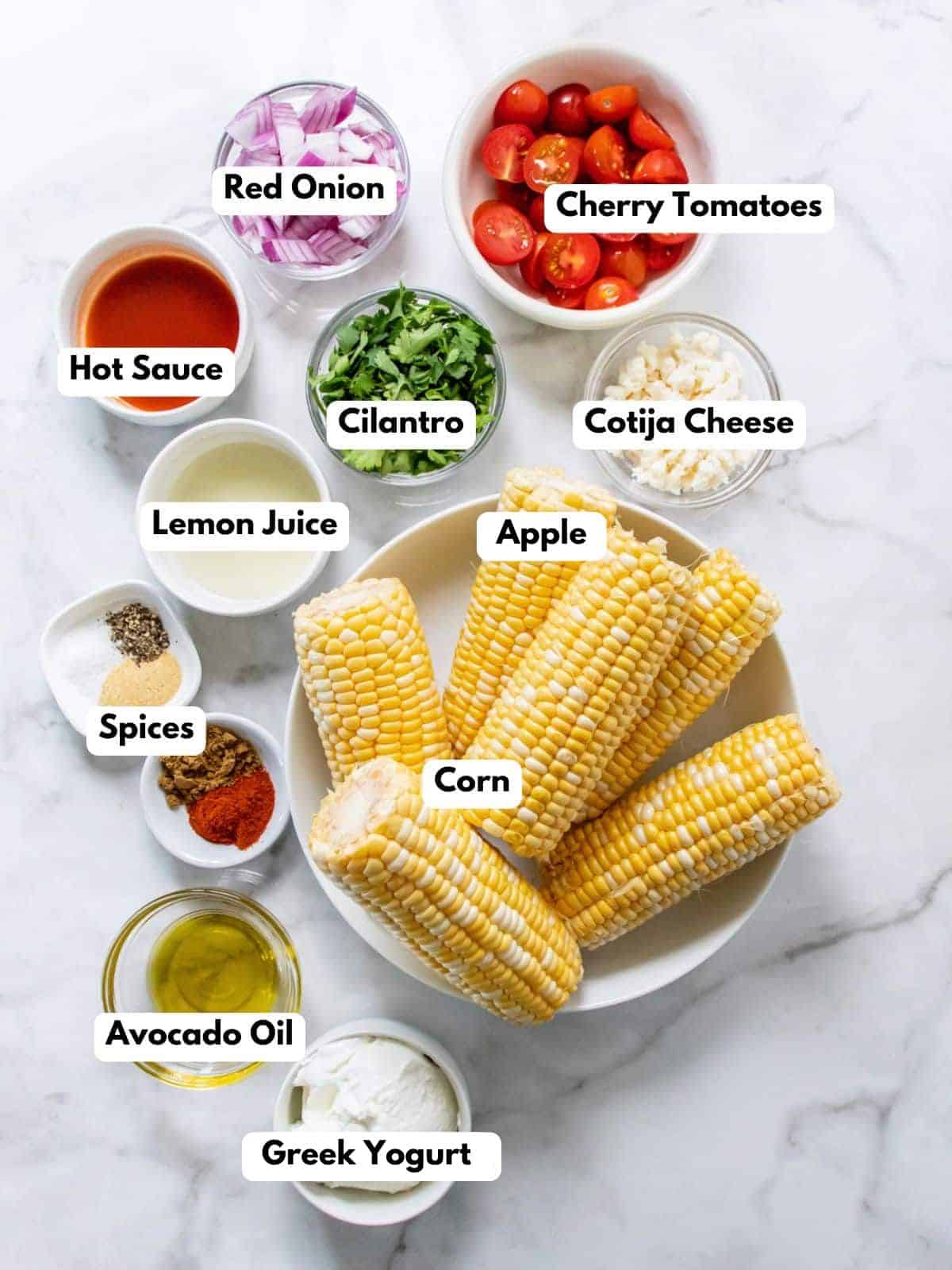 Ingredients for healthy Mexican street corn salad laid out in glass bowls on a white marbled countertop, each with a label.