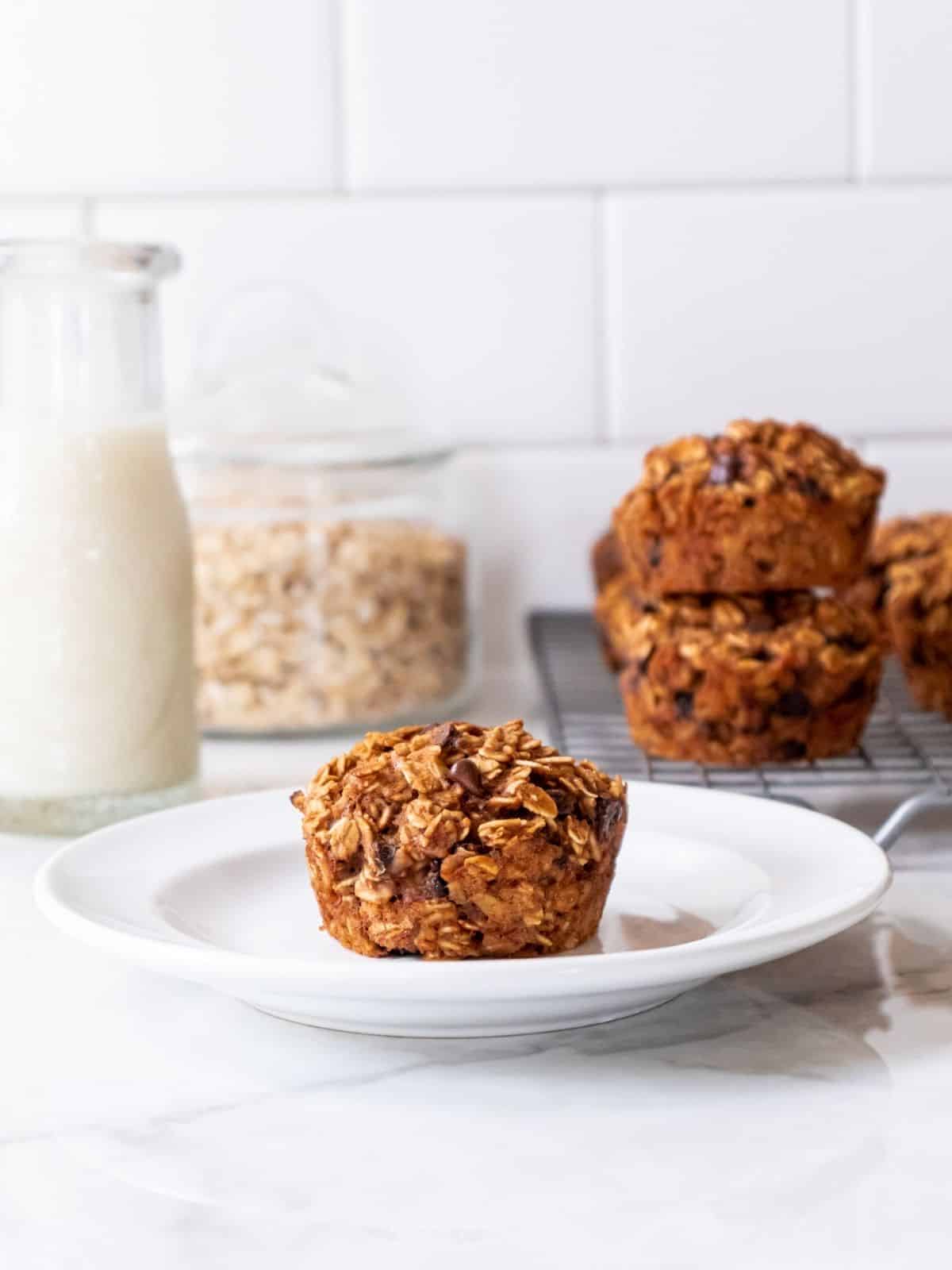 A single peanut butter banana muffin sits on a while plate. Behind there is a bottle of milk and some more muffins.