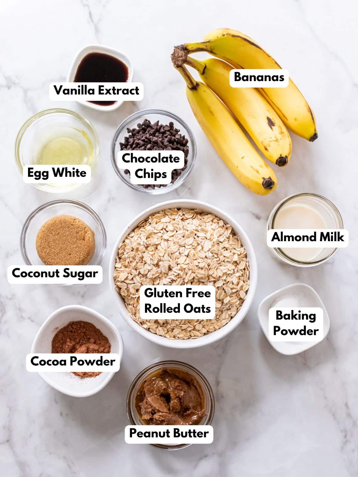 Ingredients for healthy peanut butter banana muffins presented in bowls on a white marble countertop, each one labelled. 