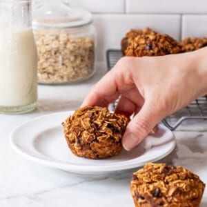 A hand picking up a peanut butter banana muffin off a white plate.