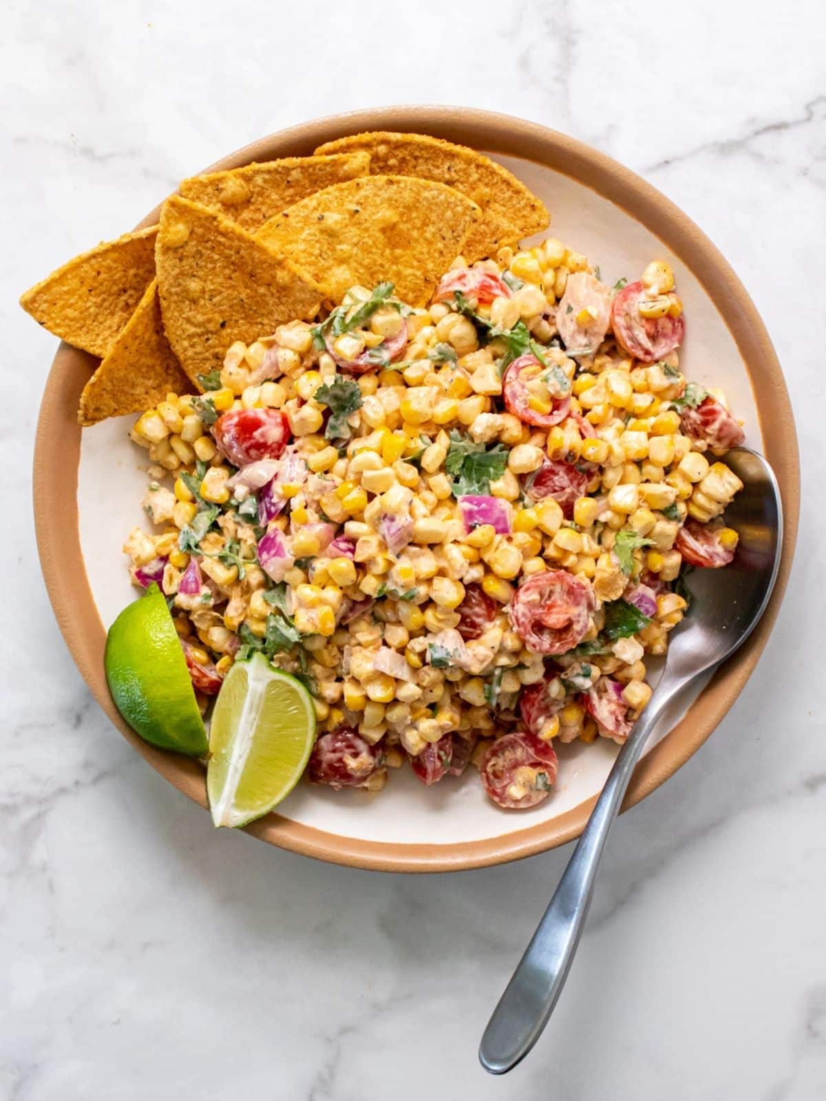 A bowl of high protein street corn with a spoon resting on the side, along with tortilla chips and lime wedges.