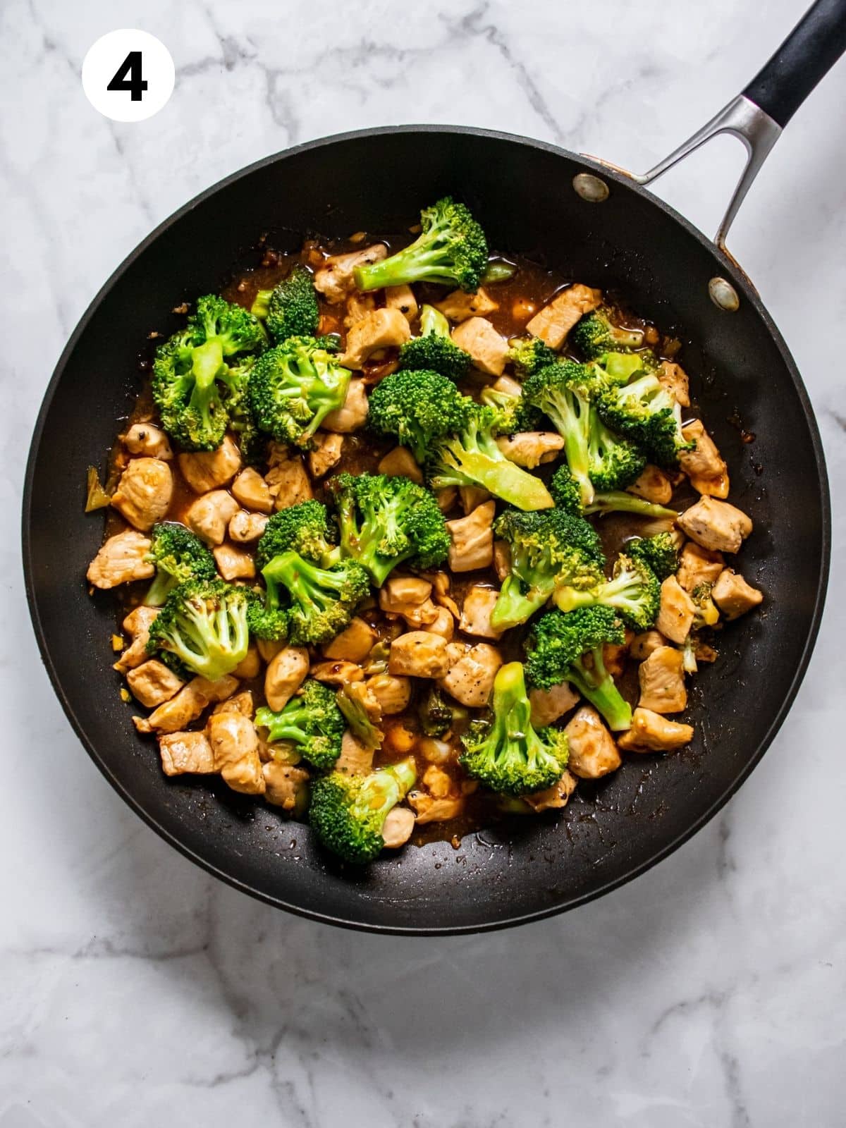 Cooked chicken and broccoli in a black skillet on a white marbled countertop.