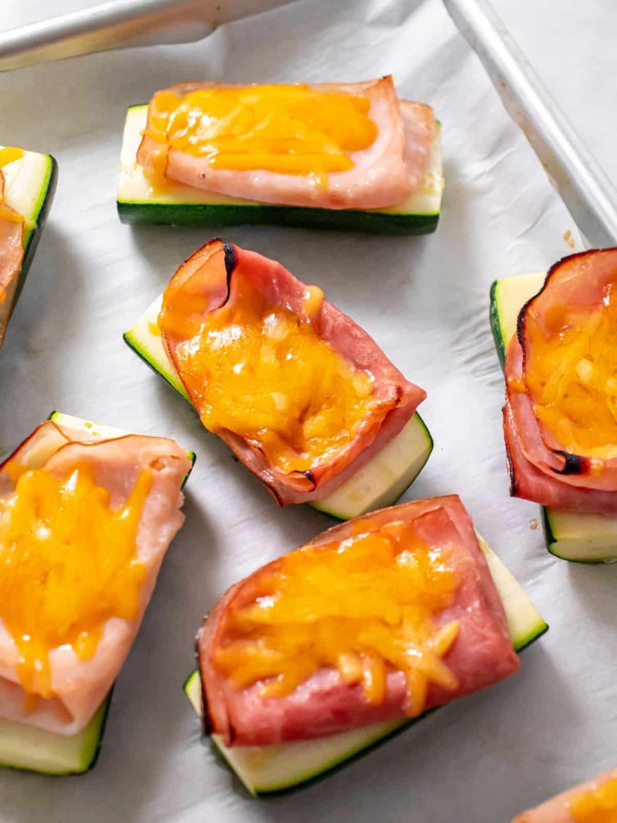 Cooked ham and turkey healthy zucchini boats topped with grated cheese sit on a foil-lined baking sheet.
