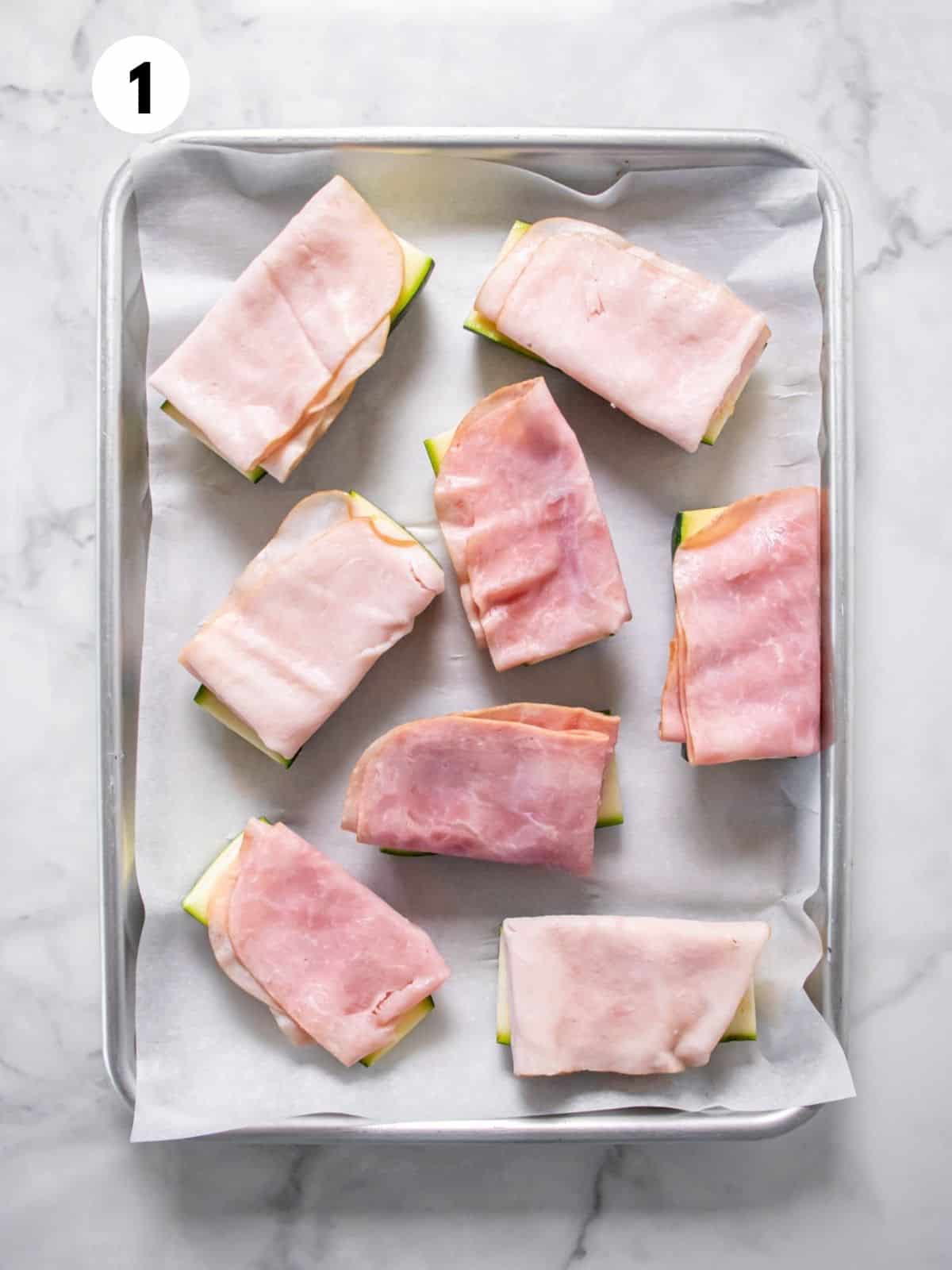 Ham and turkey-topped zucchini boats on a parchment-lined baking sheet ready for the oven.