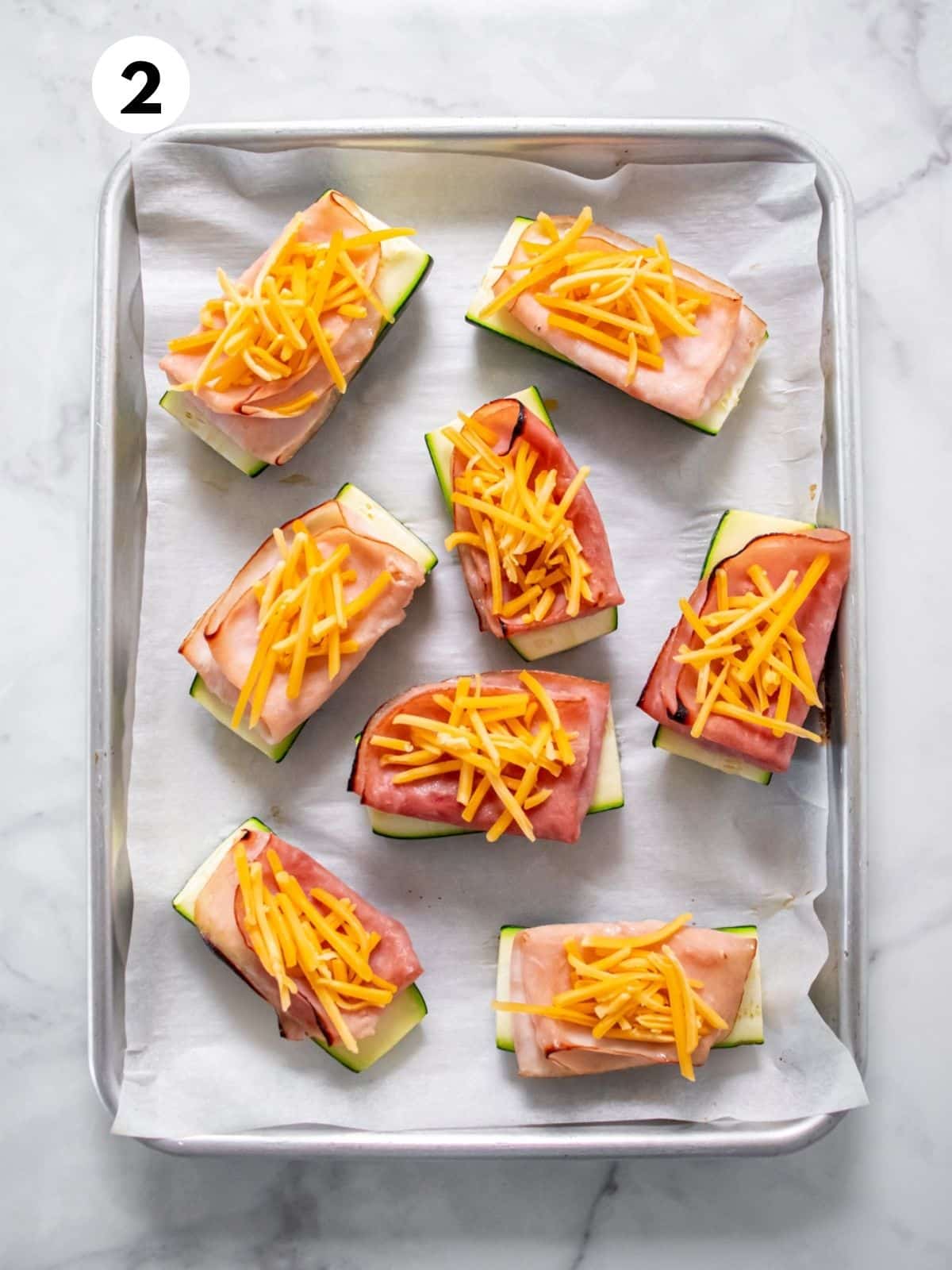 Baked ham and turkey zucchini boats topped with melted cheese, on a parchment-lined baking sheet.