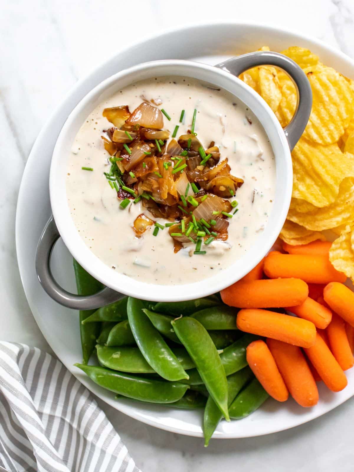 A bowl of healthy French onion dip on a plate surrounded by snap peas, carrots, and potato chips.