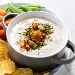 A bowl of healthy French onion dip on a plate surrounded by chips and veggies.