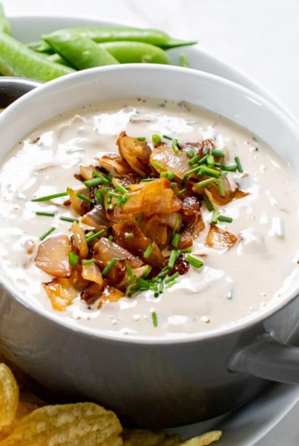 A bowl of healthy French onion dip on a plate surrounded by chips and veggies.