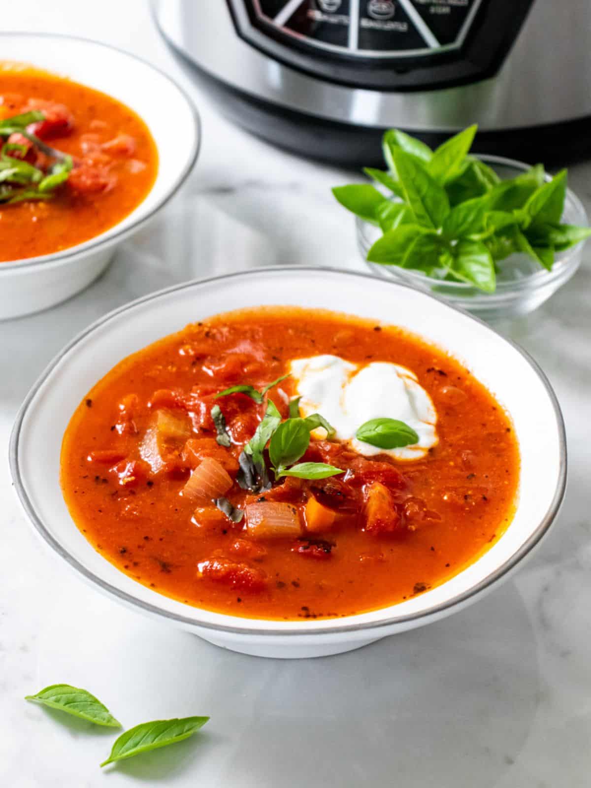 Bowls of healthy tomato soup in front of an Instant pot with a bowl of basil to the side.