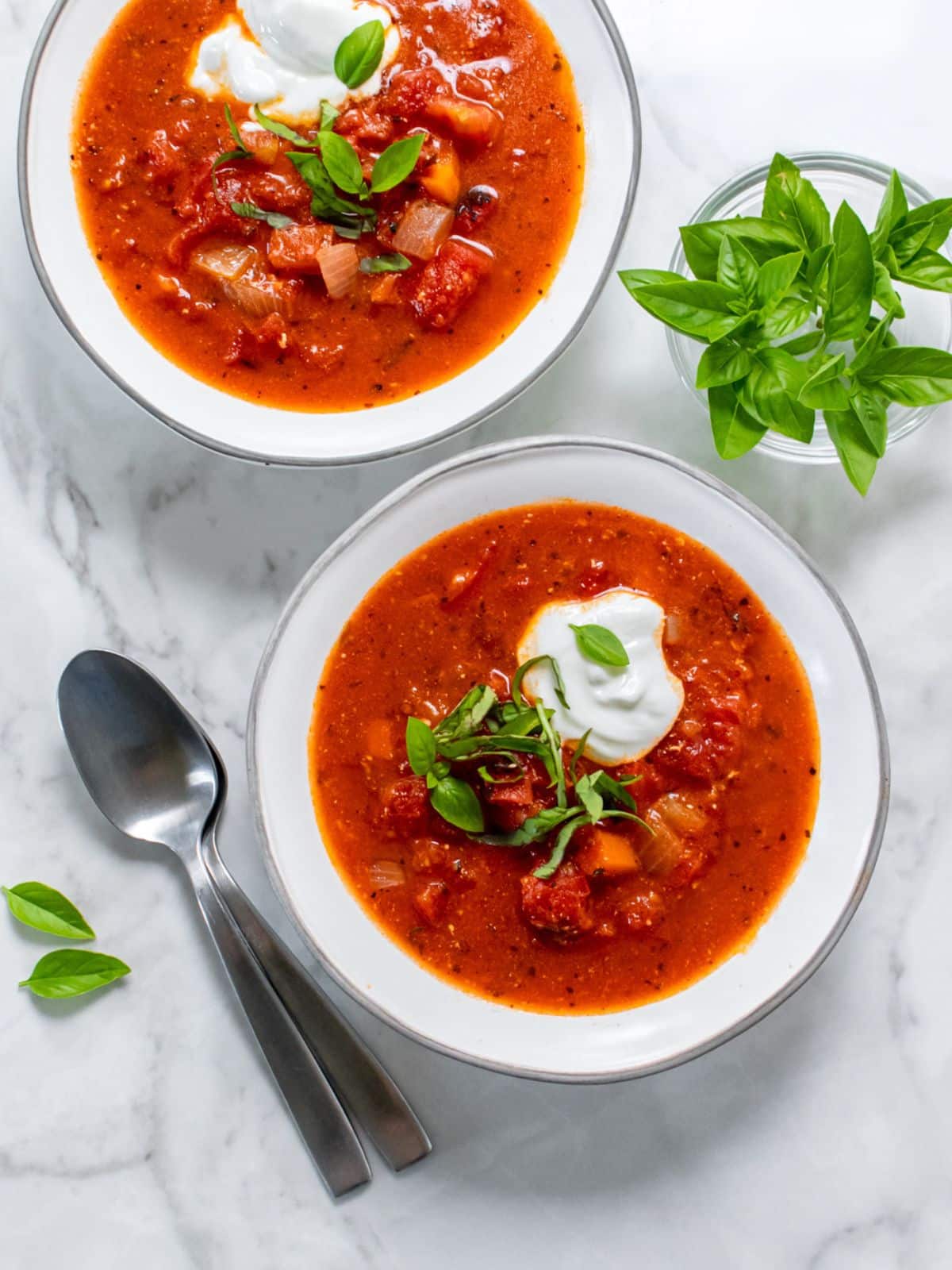 Two bowls of healthy tomato soup with a bowl of basil on a table. 