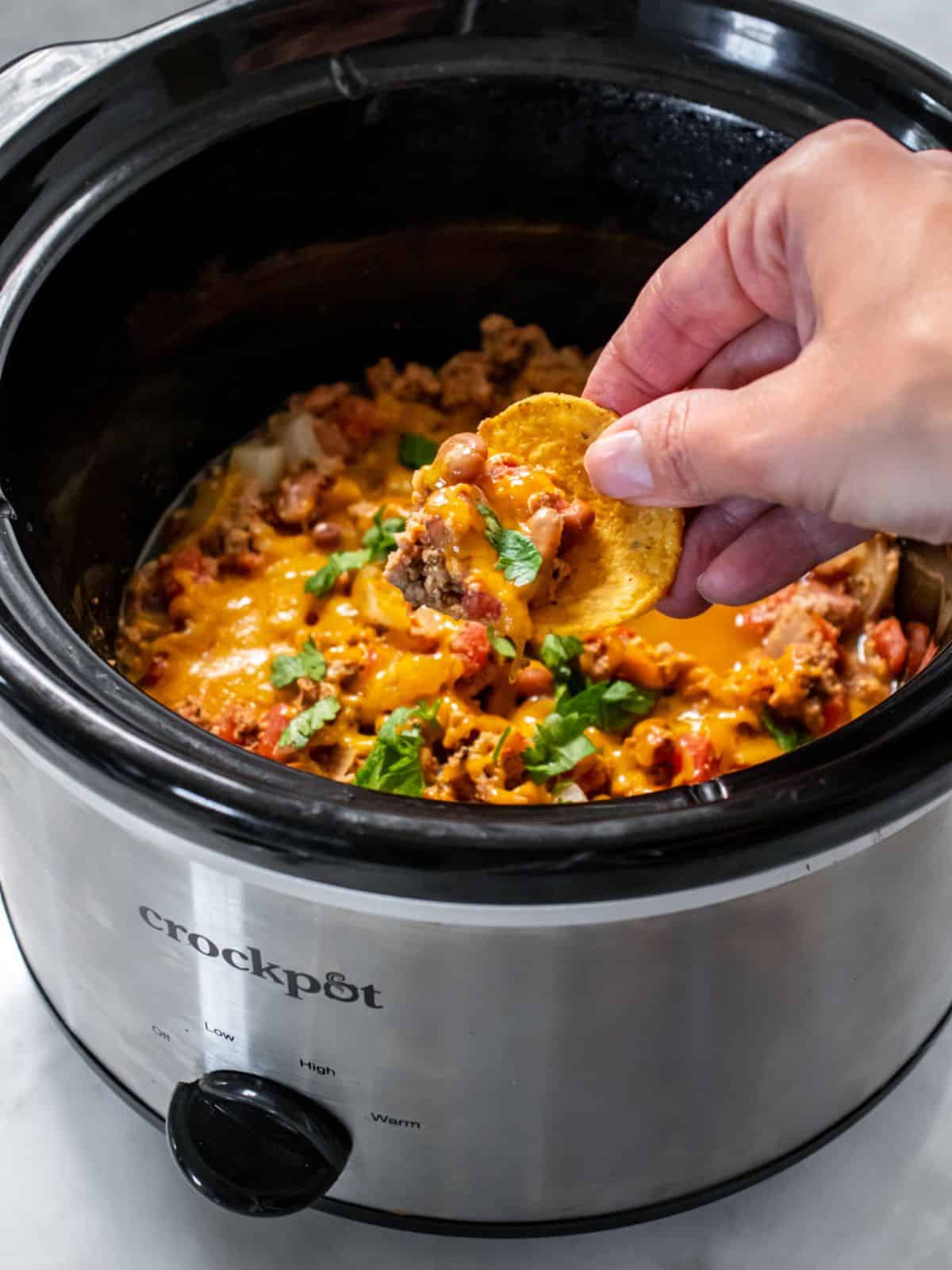A hand reaching in to dip up some crockpot chili cheese dip with a tortilla chip.