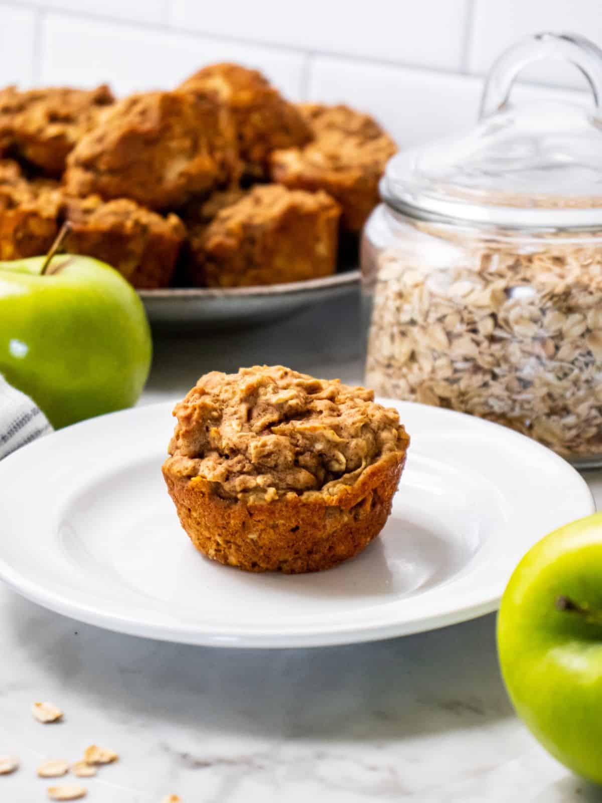 A healthy apple muffin on a plate with a green apple and a plate of more muffins in the background.