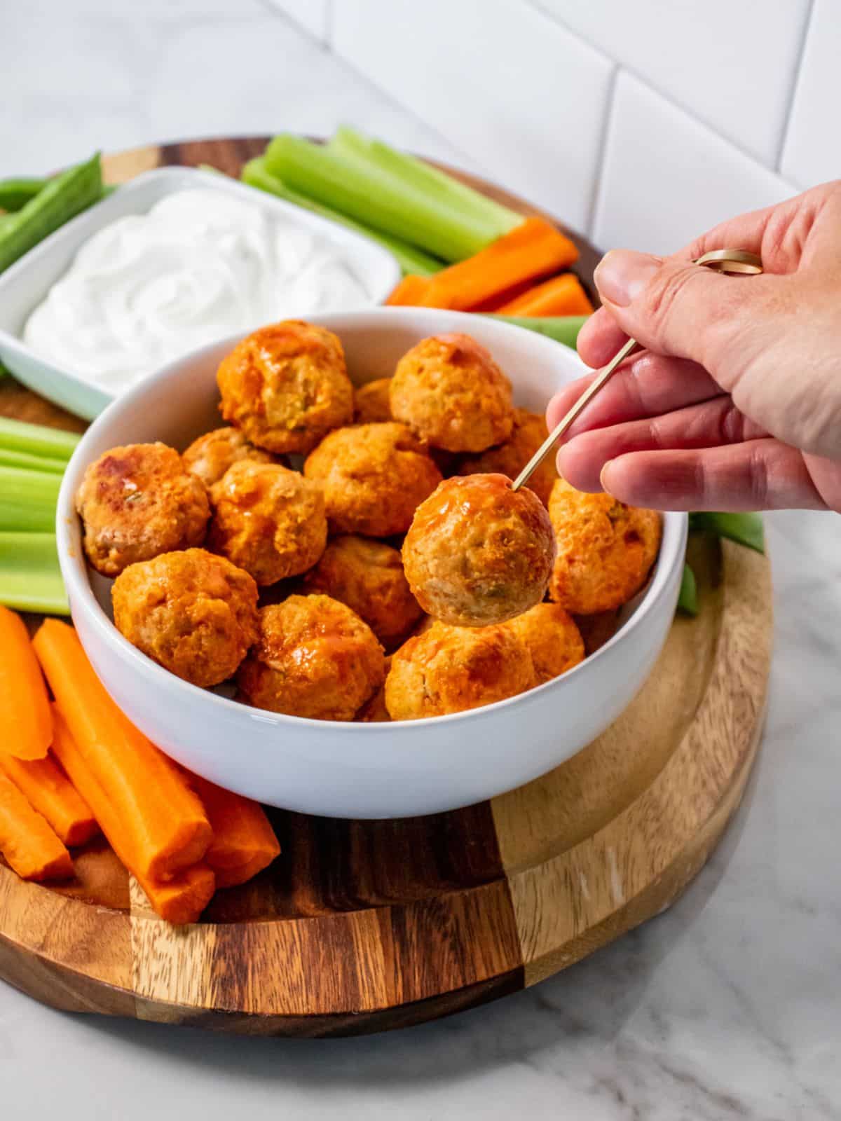 A hand using a toothpick to grab a healthy buffalo chicken meatball from a bowl on a platter.