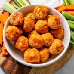 Healthy buffalo chicken meatballs in a white bowl on a round wooden cutting board.