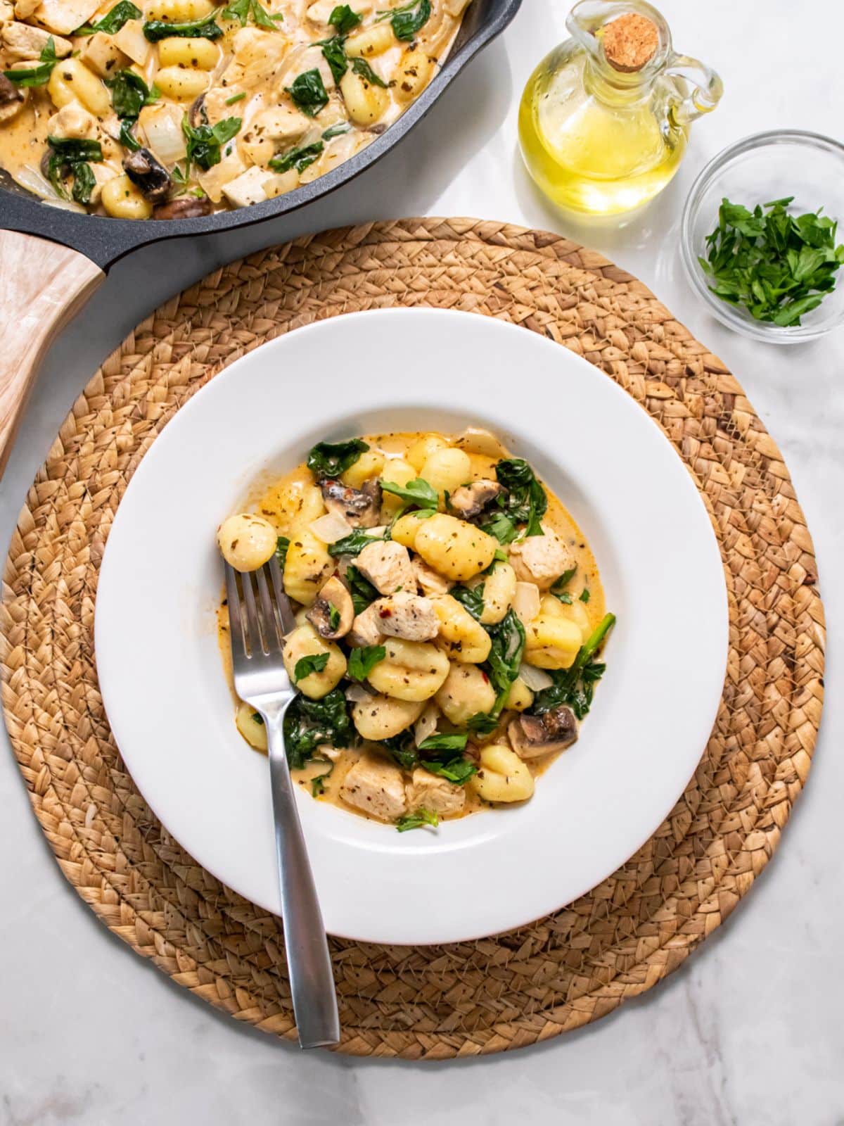 A plate of chicken and gnocchi with a fork on the dish and a skillet of more in the background.