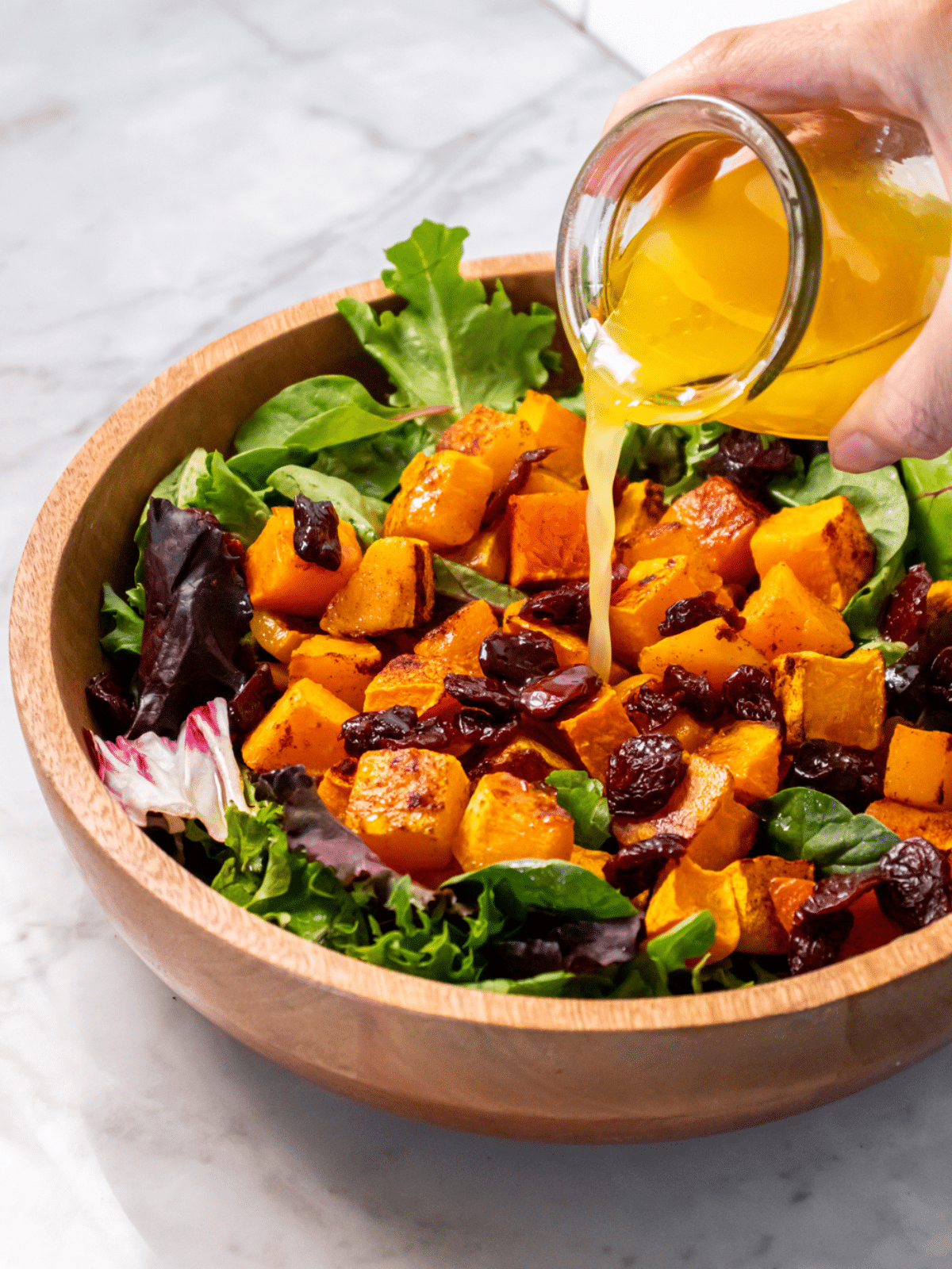 A hand pouring dressing from a glass jar over a bowl of roasted butternut squash salad.