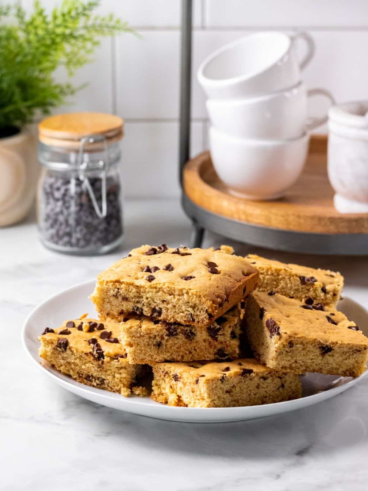 A plate of health cookie bars stacked haphazardly and a jar of chocolate chips and some coffee mugs sit at the back of the image.