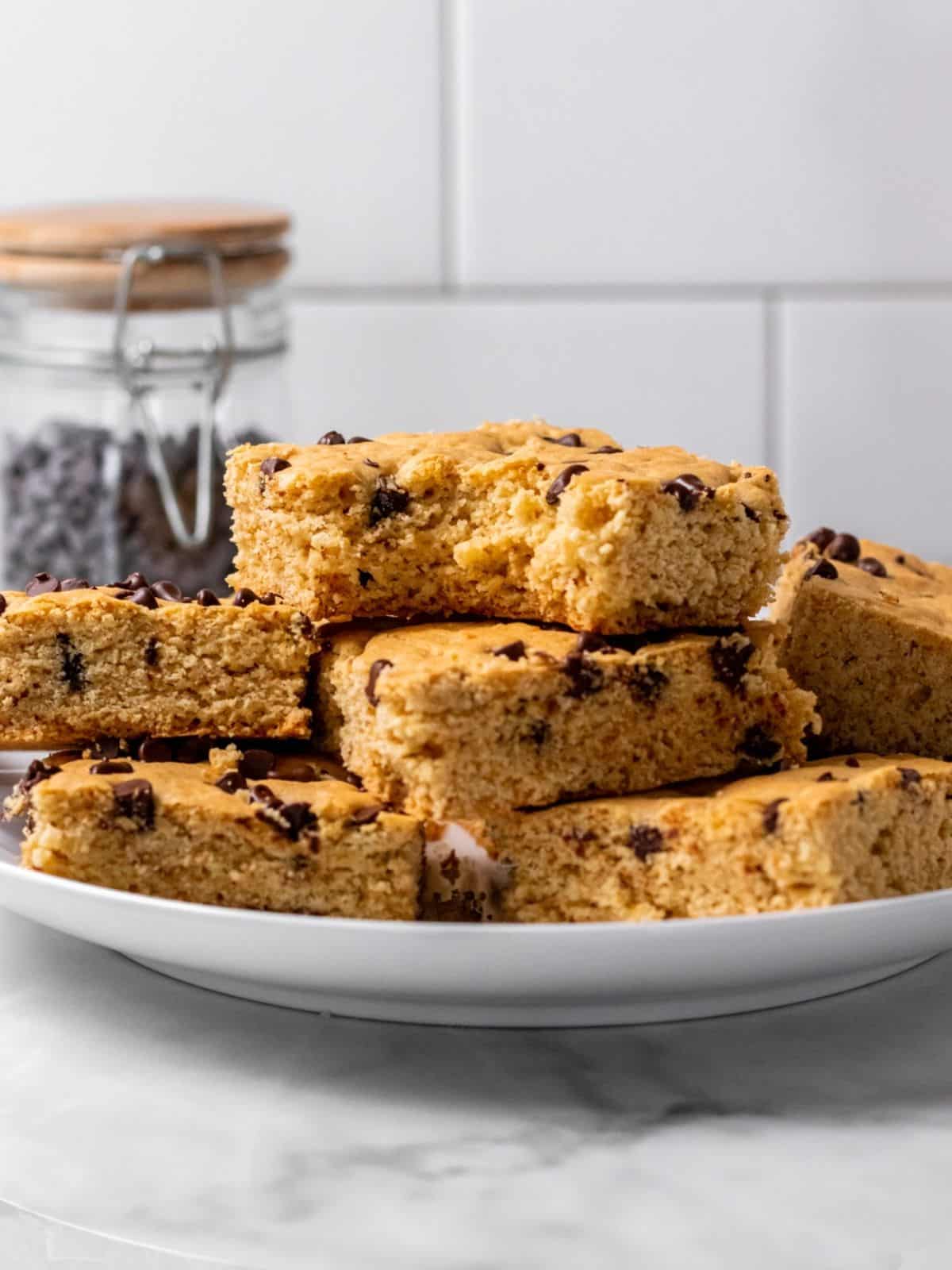 A stack of chocolate chip cookie bars on a white plate, with a jar of chocolate chips behind.