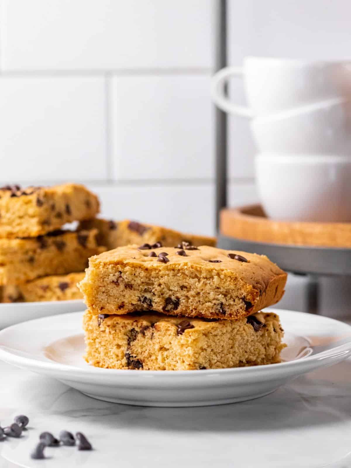 A stack of two chocolate chip cookie bars on a white plate, with more behind alongside some coffee cups.